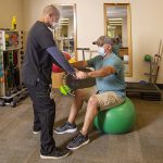 PT trainer helping man sitting on medicine ball