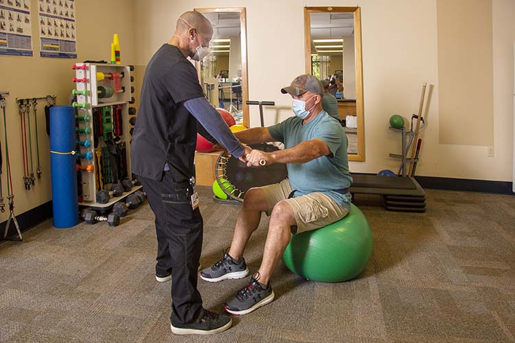 PT trainer helping man sitting on medicine ball